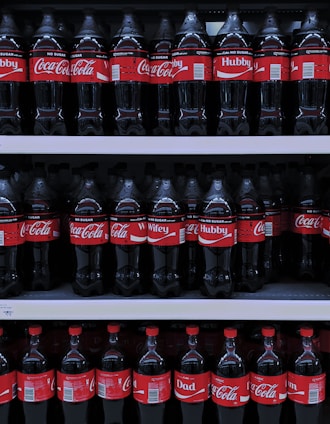 Multiple rows of Coca-Cola bottles are displayed on store shelves. The bottles have different personalized labels, including 'Hubby', 'Wifey', and 'Dad'. The red and white branding is consistent throughout.