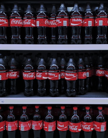 Multiple rows of Coca-Cola bottles are displayed on store shelves. The bottles have different personalized labels, including 'Hubby', 'Wifey', and 'Dad'. The red and white branding is consistent throughout.