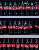 Multiple rows of Coca-Cola bottles are displayed on store shelves. The bottles have different personalized labels, including 'Hubby', 'Wifey', and 'Dad'. The red and white branding is consistent throughout.