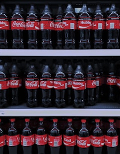 Multiple rows of Coca-Cola bottles are displayed on store shelves. The bottles have different personalized labels, including 'Hubby', 'Wifey', and 'Dad'. The red and white branding is consistent throughout.
