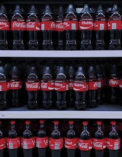 Multiple rows of Coca-Cola bottles are displayed on store shelves. The bottles have different personalized labels, including 'Hubby', 'Wifey', and 'Dad'. The red and white branding is consistent throughout.