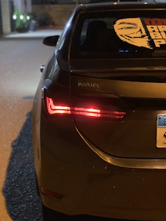 The rear of a dark-colored Toyota sedan is parked on a street at night, illuminated by the nearby streetlights. The car's left taillight is on, and a distinctive sticker featuring a stylized face is displayed on the rear windshield. A portion of a license plate is visible.