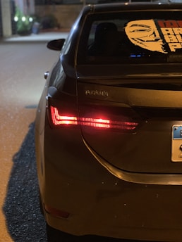 The rear of a dark-colored Toyota sedan is parked on a street at night, illuminated by the nearby streetlights. The car's left taillight is on, and a distinctive sticker featuring a stylized face is displayed on the rear windshield. A portion of a license plate is visible.