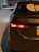 The rear of a dark-colored Toyota sedan is parked on a street at night, illuminated by the nearby streetlights. The car's left taillight is on, and a distinctive sticker featuring a stylized face is displayed on the rear windshield. A portion of a license plate is visible.