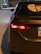 The rear of a dark-colored Toyota sedan is parked on a street at night, illuminated by the nearby streetlights. The car's left taillight is on, and a distinctive sticker featuring a stylized face is displayed on the rear windshield. A portion of a license plate is visible.