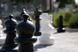 An outdoor giant chess set with prominently featured black and white pawns and other pieces on a concrete surface. The background includes blurred greenery and a building, suggesting a garden or park setting.