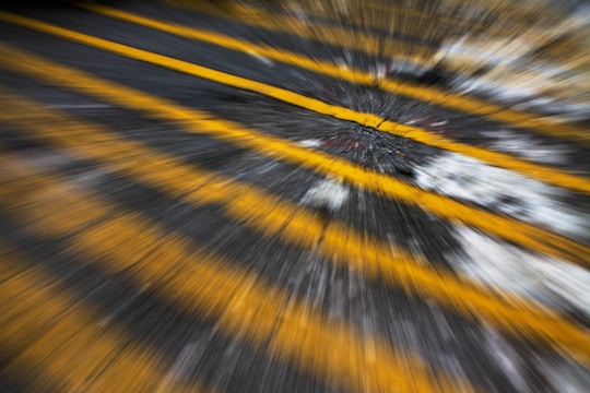 A dynamic, motion-blurred close-up of road markings. Yellow stripes are prominently featured, with a cracked, weathered asphalt background. The image gives an impression of speed and movement.