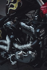 A close-up view of a motorcycle engine, featuring metallic components and wrapped exhaust pipes. The metal surfaces display a shiny and reflective quality, while decorative flame motifs in yellow and red colors are visible on the side of the vehicle.
