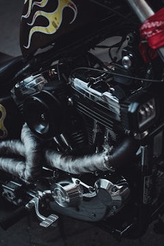 A close-up view of a motorcycle engine, featuring metallic components and wrapped exhaust pipes. The metal surfaces display a shiny and reflective quality, while decorative flame motifs in yellow and red colors are visible on the side of the vehicle.