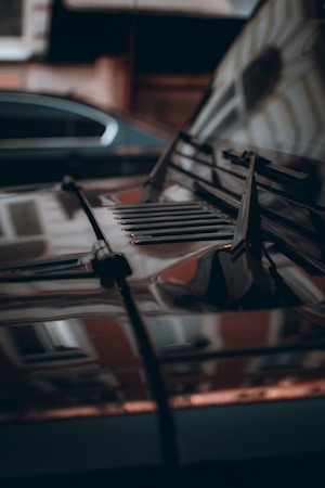 A close-up view of a car hood with reflections of nearby buildings and another vehicle in the glossy surface. The image features parts of the windshield wipers and air vent slats, suggesting an urban environment with a focus on the intricate details of the car's design.