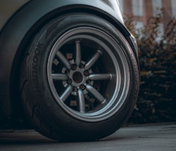 A close-up image of a car wheel showcasing the tire and metallic rim in detail. The tire brand 'Hankook' is visible, and the wheel appears sturdy and well-maintained. The background includes blurred foliage and a pavement surface.