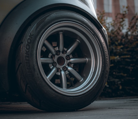 A close-up image of a car wheel showcasing the tire and metallic rim in detail. The tire brand 'Hankook' is visible, and the wheel appears sturdy and well-maintained. The background includes blurred foliage and a pavement surface.