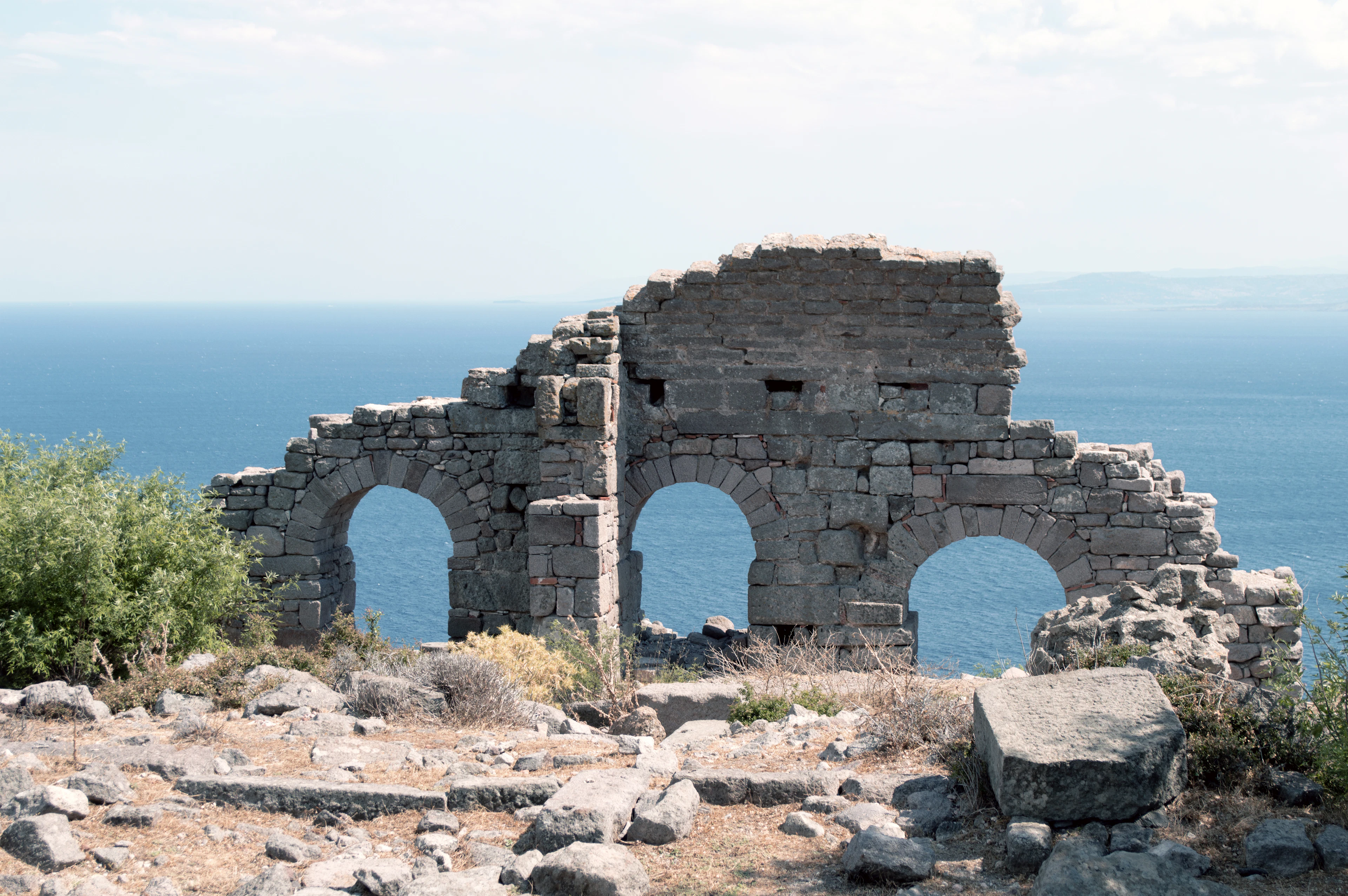Ancient stone arches on a hillside above the Aegean, with an island on the horizon