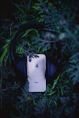 A smartphone with a visible Apple logo rests on a pair of black headphones surrounded by lush green foliage. The scene creates a contrast between technology and nature, as the modern devices are partially hidden among the vibrant leaves.