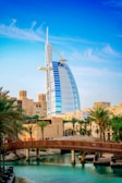 A modern sail-shaped skyscraper stands prominently among palm trees and traditional Middle Eastern architecture with a wooden bridge spanning a serene body of water in the foreground. The sky above is clear and blue with wispy clouds.