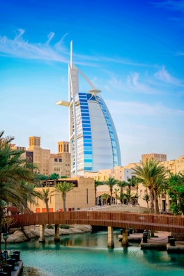 A modern sail-shaped skyscraper stands prominently among palm trees and traditional Middle Eastern architecture with a wooden bridge spanning a serene body of water in the foreground. The sky above is clear and blue with wispy clouds.