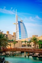 A modern sail-shaped skyscraper stands prominently among palm trees and traditional Middle Eastern architecture with a wooden bridge spanning a serene body of water in the foreground. The sky above is clear and blue with wispy clouds.