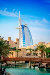 A modern sail-shaped skyscraper stands prominently among palm trees and traditional Middle Eastern architecture with a wooden bridge spanning a serene body of water in the foreground. The sky above is clear and blue with wispy clouds.