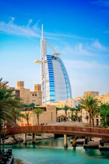 A modern sail-shaped skyscraper stands prominently among palm trees and traditional Middle Eastern architecture with a wooden bridge spanning a serene body of water in the foreground. The sky above is clear and blue with wispy clouds.