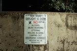 A worn concrete wall displays a sign listing park regulations, including operation hours from daylight to dusk and prohibitions against alcohol, skateboarding, littering, rollerblading, camping, and disorderly conduct. The sign emphasizes that violators will face prosecution.