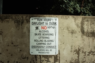 A worn concrete wall displays a sign listing park regulations, including operation hours from daylight to dusk and prohibitions against alcohol, skateboarding, littering, rollerblading, camping, and disorderly conduct. The sign emphasizes that violators will face prosecution.