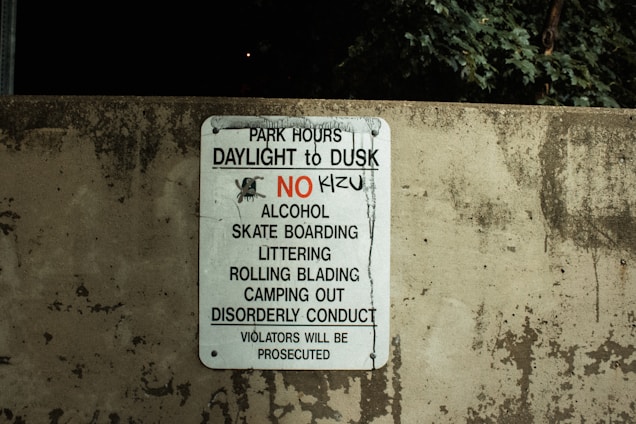 A worn concrete wall displays a sign listing park regulations, including operation hours from daylight to dusk and prohibitions against alcohol, skateboarding, littering, rollerblading, camping, and disorderly conduct. The sign emphasizes that violators will face prosecution.