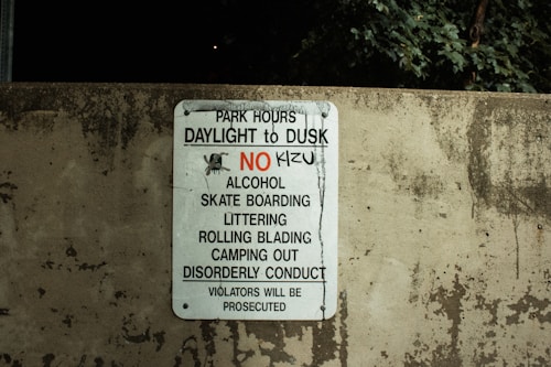 A worn concrete wall displays a sign listing park regulations, including operation hours from daylight to dusk and prohibitions against alcohol, skateboarding, littering, rollerblading, camping, and disorderly conduct. The sign emphasizes that violators will face prosecution.