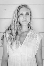 A black and white portrait of a person with long, wavy hair wearing a sleeveless, lacy white top. They are standing against a plain, light-colored wooden wall, looking slightly to the side.