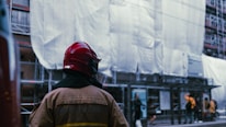 A firefighter wearing a red helmet and brown protective jacket stands in front of a building covered in scaffolding and white sheeting. Several people are visible in the background, including some standing and talking near a bus stop.
