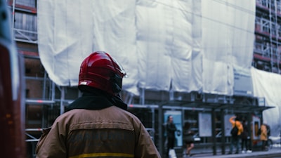 A firefighter wearing a red helmet and brown protective jacket stands in front of a building covered in scaffolding and white sheeting. Several people are visible in the background, including some standing and talking near a bus stop.