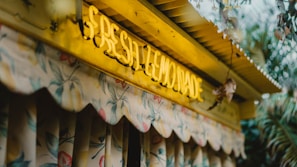 A vibrant yellow neon sign displaying the words 'Fresh Lemonade' is mounted on a wooden structure. The building features a decorative fabric awning with a floral pattern in pastel hues. Surrounding the structure are green leafy plants, adding to the fresh and natural ambiance.