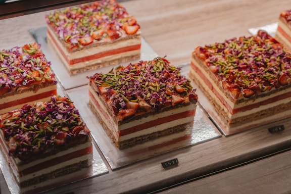 A series of square, layered cakes are displayed on a countertop. Each cake has alternating layers of cream and red filling, topped with a colorful mix of finely chopped green pistachios and red flower petals. The cakes are placed on square silver plates in a well-lit environment.