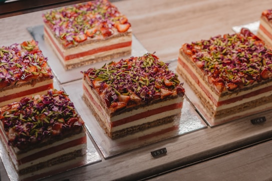 A series of square, layered cakes are displayed on a countertop. Each cake has alternating layers of cream and red filling, topped with a colorful mix of finely chopped green pistachios and red flower petals. The cakes are placed on square silver plates in a well-lit environment.