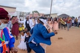 A vibrant outdoor celebration featuring a group of people dressed in formal and traditional attire. A man in a blue suit appears to be dancing, holding a stick. Several bridesmaids in peach dresses stand nearby, while children in formal wear are also present. A brass band is playing, creating a lively atmosphere. Onlookers surround the scene, capturing the moment with their phones.
