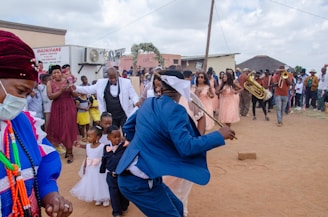 A vibrant outdoor celebration featuring a group of people dressed in formal and traditional attire. A man in a blue suit appears to be dancing, holding a stick. Several bridesmaids in peach dresses stand nearby, while children in formal wear are also present. A brass band is playing, creating a lively atmosphere. Onlookers surround the scene, capturing the moment with their phones.