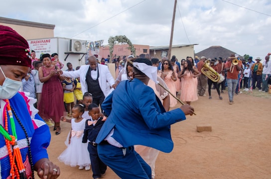 A vibrant outdoor celebration featuring a group of people dressed in formal and traditional attire. A man in a blue suit appears to be dancing, holding a stick. Several bridesmaids in peach dresses stand nearby, while children in formal wear are also present. A brass band is playing, creating a lively atmosphere. Onlookers surround the scene, capturing the moment with their phones.