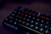 A close-up of a mechanical keyboard with illuminated keys. The keys have a backlight featuring predominantly red and blue colors, creating a contrast against the dark background. The keyboard is set in a dimly lit environment, highlighting the glowing effect of the keys.