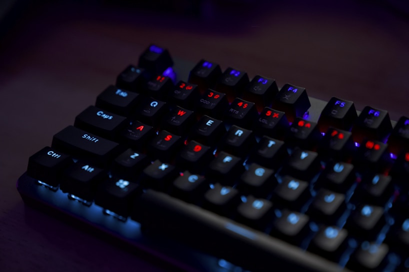 A close-up of a mechanical keyboard with illuminated keys. The keys have a backlight featuring predominantly red and blue colors, creating a contrast against the dark background. The keyboard is set in a dimly lit environment, highlighting the glowing effect of the keys.
