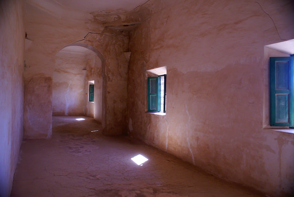 The image depicts a room with a large arched doorway on one side, flanked by two windows with green shutters and white curtains. The walls are made of light-colored stone or concrete, giving the space an old-world charm. A single beam of sunlight pierces through the window in the center of the room, casting a warm glow that illuminates the scene.