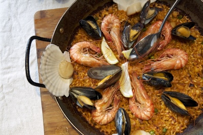 A close-up view of a paella dish in a round, dark pan. The rice is cooked to a golden brown and is garnished with large shrimp, mussels, slices of lemon, and a scallop shell. The shrimp are arranged in a circular pattern, with their tails pointing outward. The mussels are open, revealing their orange centers. The dish rests on a wooden board, partially covered by a white tablecloth.