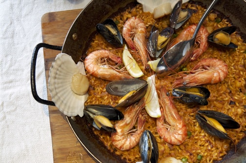 A close-up view of a paella dish in a round, dark pan. The rice is cooked to a golden brown and is garnished with large shrimp, mussels, slices of lemon, and a scallop shell. The shrimp are arranged in a circular pattern, with their tails pointing outward. The mussels are open, revealing their orange centers. The dish rests on a wooden board, partially covered by a white tablecloth.