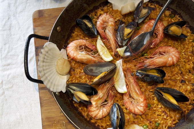 A close-up view of a paella dish in a round, dark pan. The rice is cooked to a golden brown and is garnished with large shrimp, mussels, slices of lemon, and a scallop shell. The shrimp are arranged in a circular pattern, with their tails pointing outward. The mussels are open, revealing their orange centers. The dish rests on a wooden board, partially covered by a white tablecloth.