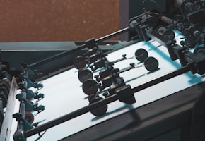 A close-up view of a printing press machine with rollers and sheets of paper. Various mechanical parts including wheels and levers are visible, emphasizing the industrial nature of the machinery.