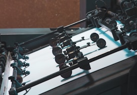 A close-up view of a printing press machine with rollers and sheets of paper. Various mechanical parts including wheels and levers are visible, emphasizing the industrial nature of the machinery.