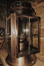 A vintage metal lantern with glass panels rests on a wooden surface. The lantern has a weathered, bronze finish and intricate detailing. It is placed against a stone wall, adding an old-world charm to the setting.