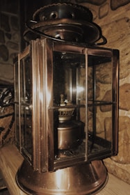 A vintage metal lantern with glass panels rests on a wooden surface. The lantern has a weathered, bronze finish and intricate detailing. It is placed against a stone wall, adding an old-world charm to the setting.