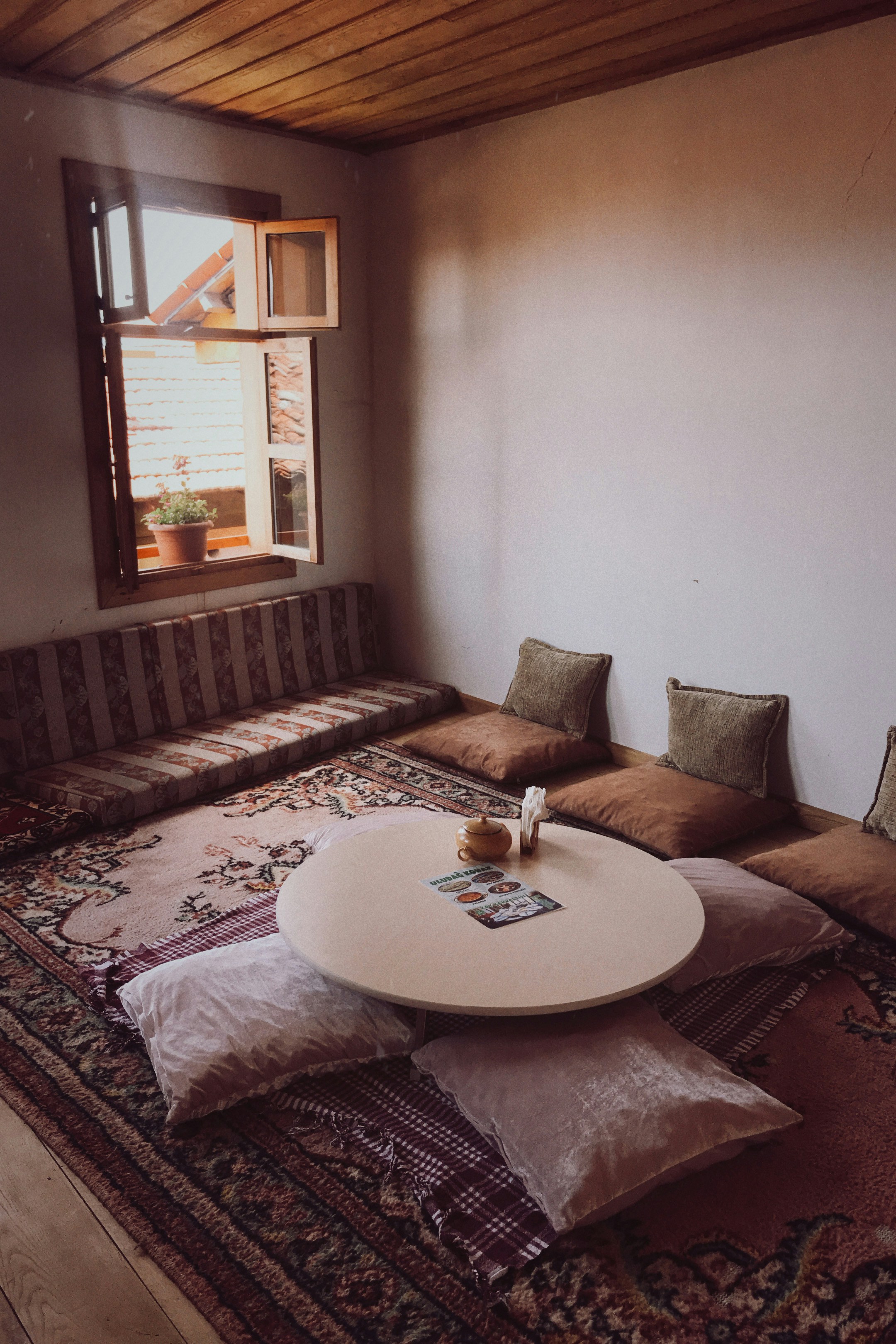 A cozy room with a low round table surrounded by soft cushions and pillows on a patterned rug. An open window with a view of rooftops lets in natural light, enhancing the warm wooden ceiling and neutral walls. A small plant sits on the window ledge, adding a touch of greenery.