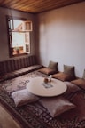 A cozy room with a low round table surrounded by soft cushions and pillows on a patterned rug. An open window with a view of rooftops lets in natural light, enhancing the warm wooden ceiling and neutral walls. A small plant sits on the window ledge, adding a touch of greenery.