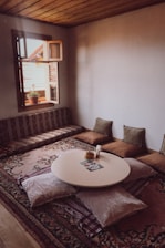 A cozy room with a low round table surrounded by soft cushions and pillows on a patterned rug. An open window with a view of rooftops lets in natural light, enhancing the warm wooden ceiling and neutral walls. A small plant sits on the window ledge, adding a touch of greenery.