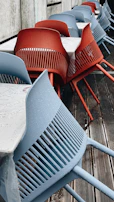 A row of modern chairs, some in blue and some in red, are aligned next to tables with white tablecloths. The chairs have a sleek design with slatted backs and are placed on a wooden deck. Water droplets are visible on the chairs, indicating recent rain.